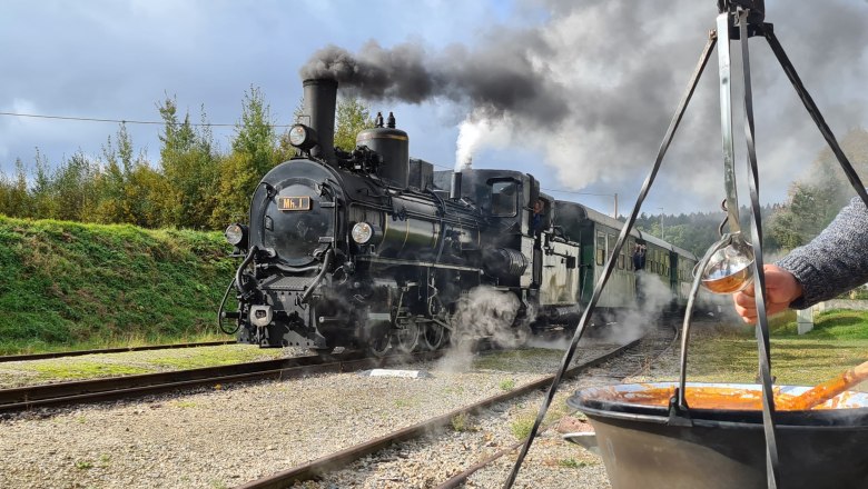 Gulaschzug, © NÖVOG/Miedler Eine Dampflokomotive fährt auf Schienen, während im Vordergrund ein Kessel mit Gulasch über einem Feuer hängt.
