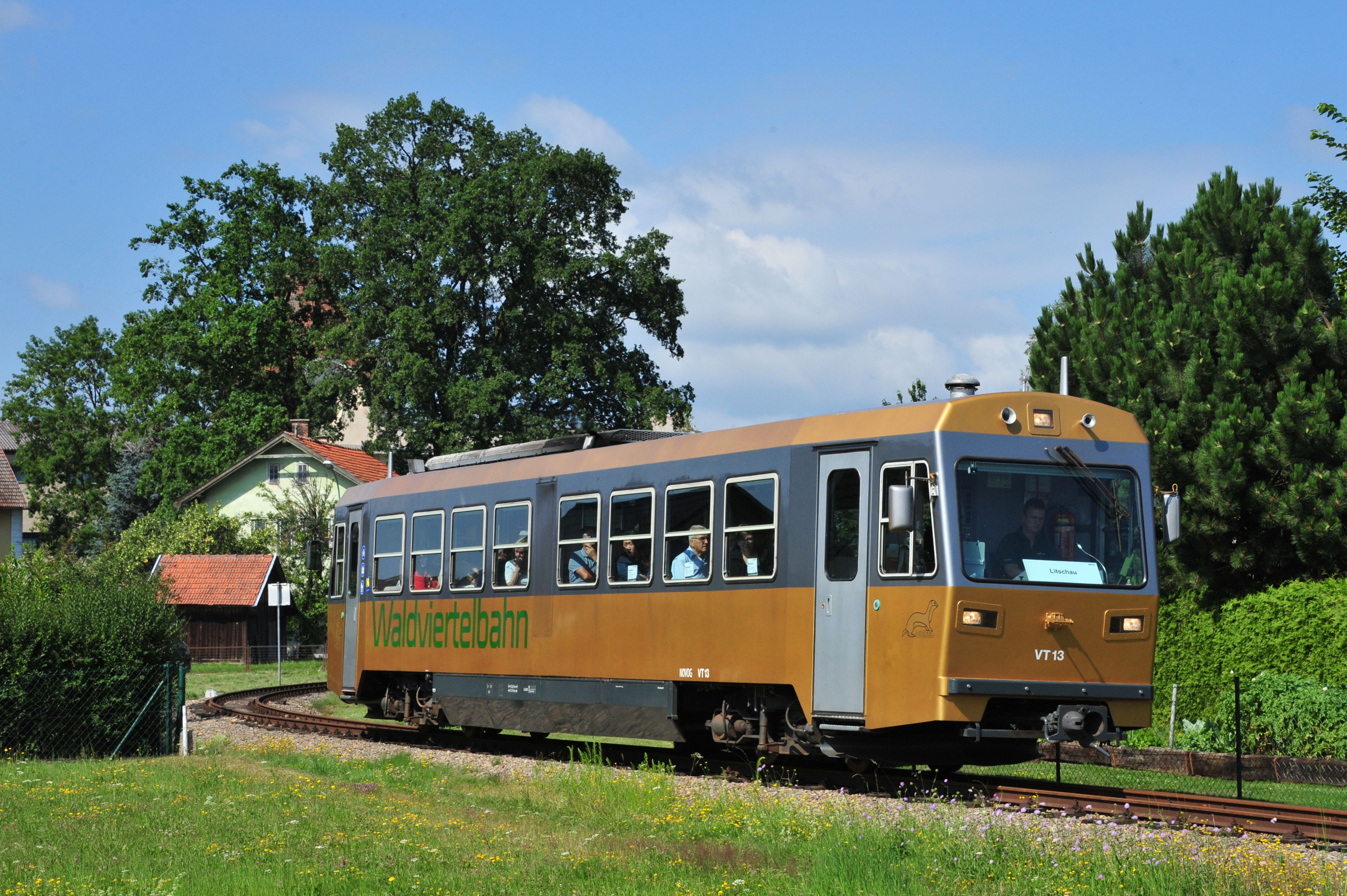 Goldfarbener Triebwagen der Waldviertelbahn fährt durch eine Kurve. Fahrgäste sitzen im Zug; im Hintergrund stehen Häuser und Bäume.