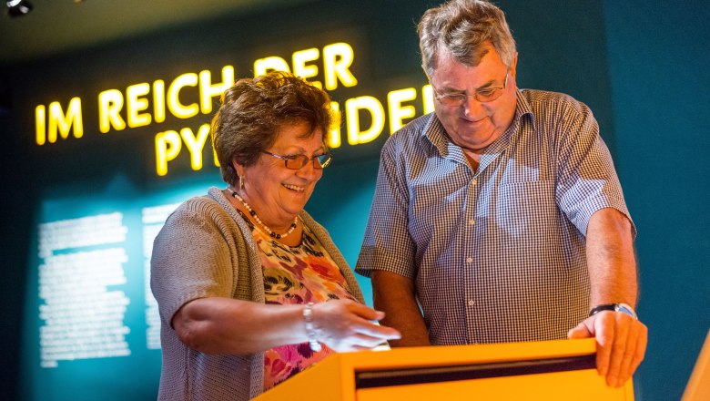 Zwei Personen betrachten ein interaktives Display in einem Museum mit der Aufschrift 'Im Reich der Pyramiden' im Hintergrund.