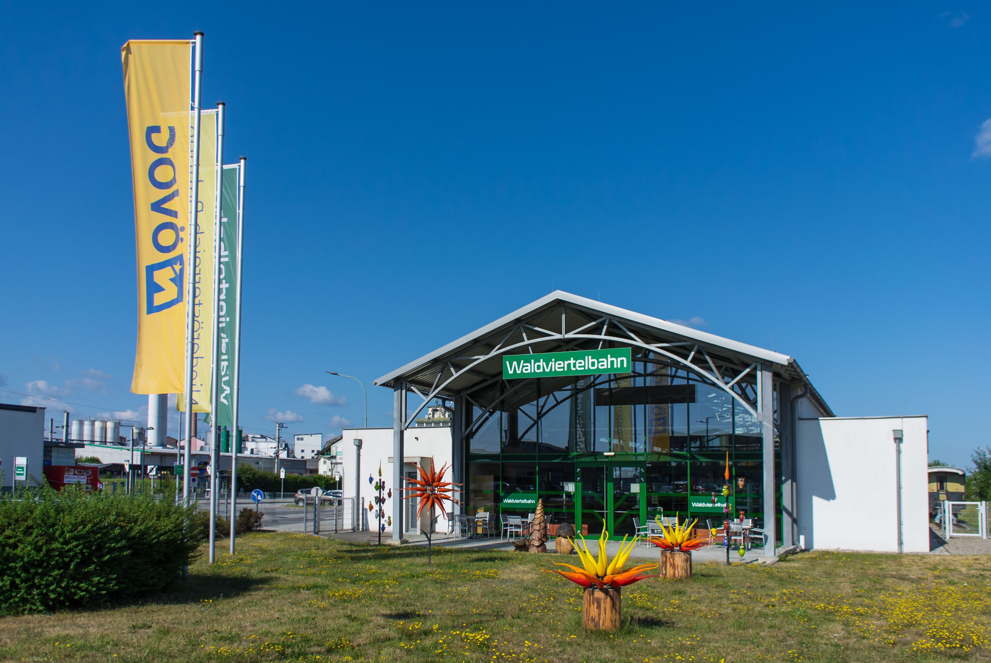 Gebäude der Waldviertelbahn in Gmünd mit Glasfassade, Fahnen und farbigen Glasskulpturen auf der Grünfläche davor.