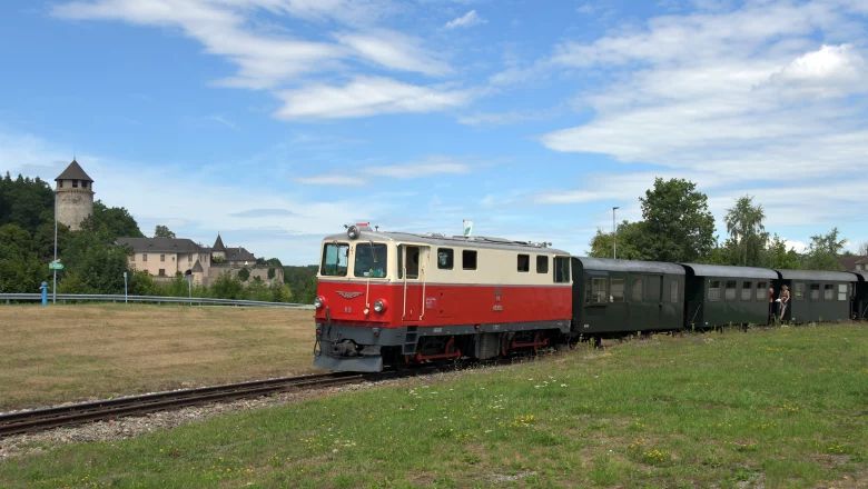Rot-beige Diesellokomotive mit dunklen Passagierwaggons auf einer eingleisigen Bahnstrecke vor einer Schlossanlage.