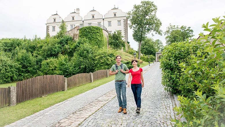 Zwei Personen mit Rucksäcken gehen auf einem gepflasterten Weg unterhalb eines hellen Schlossgebäudes bergab.