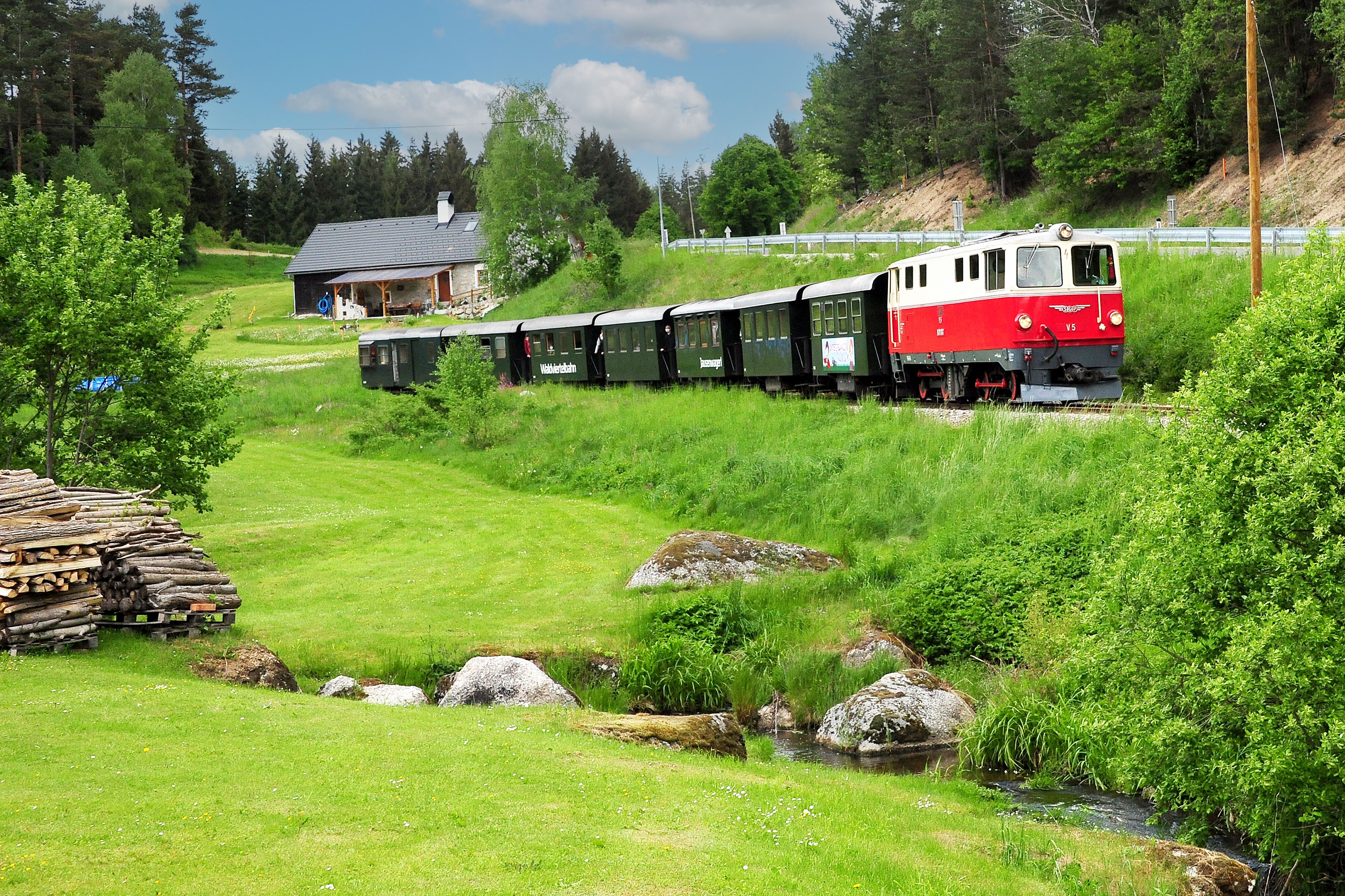 Rote Diesellokomotive zieht mehrere grüne Waggons durch eine hügelige Landschaft mit Wiesen, Bäumen, einem Bach und einem Haus.