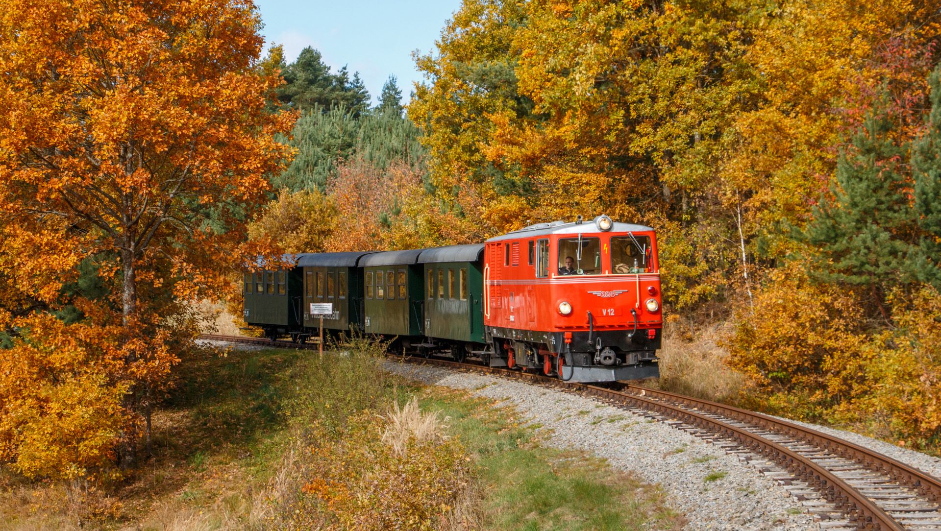 Ein roter Zug fährt durch eine herbstliche Landschaft mit bunten Bäumen.