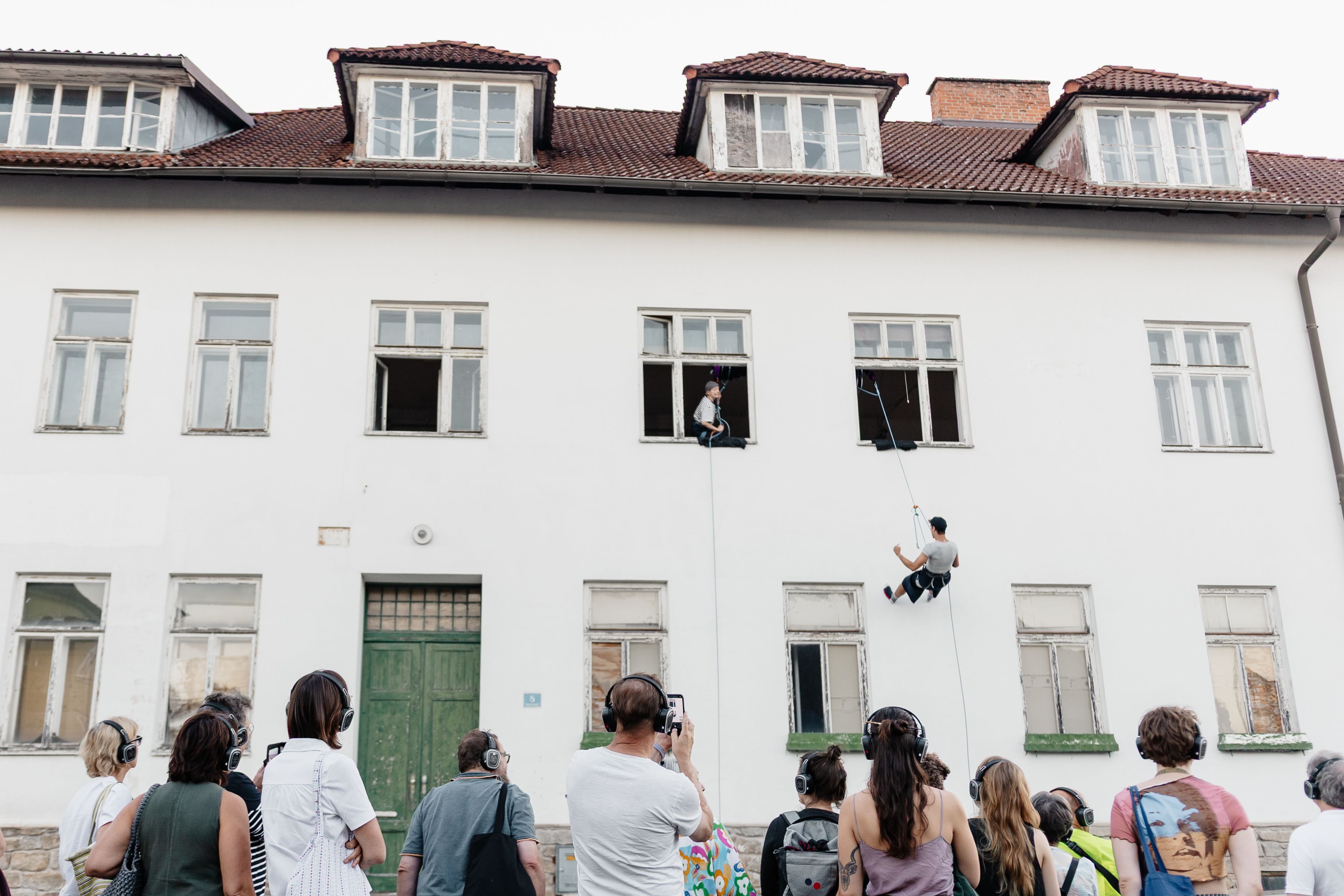 Menschen mit Kopfhörern beobachten eine Performance an einer weißen Hausfassade; zwei Personen hängen an Seilen in den Fenstern.