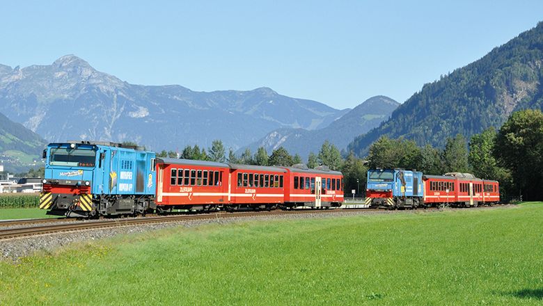Zwei blau-rote Züge der Zillertalbahn fahren durch eine grüne Wiesenlandschaft. Im Hintergrund erheben sich bewaldete Hänge und markante Berggipfel.