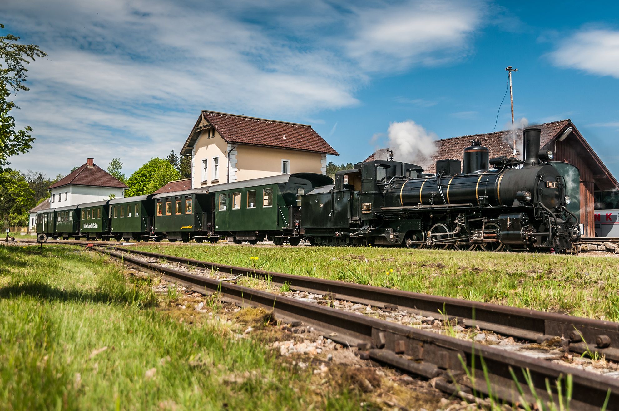 Schwarze Dampflokomotive mit grünen Personenwagen steht auf einem Gleis vor mehreren Gebäuden. Aus dem Schornstein steigt Dampf auf.