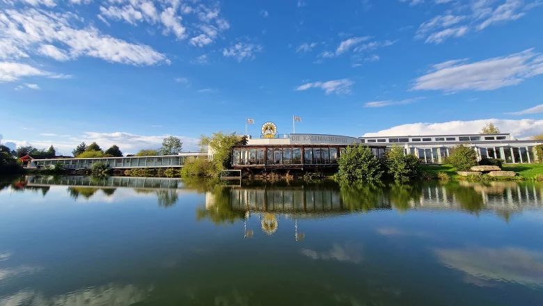 Langgestrecktes Gebäude mit Verglasung am Ufer eines Teiches, in dem sich das Bauwerk und der blaue Himmel spiegeln.