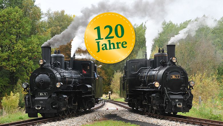 Zwei historische Dampflokomotiven auf einem Gleisdreieck in einer ländlichen Umgebung mit einem Schild '120 Jahre'.