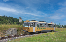 Ein goldener Zug der Waldviertelbahn f&auml;hrt durch eine gr&uuml;ne Landschaft mit einem Turm im Hintergrund.