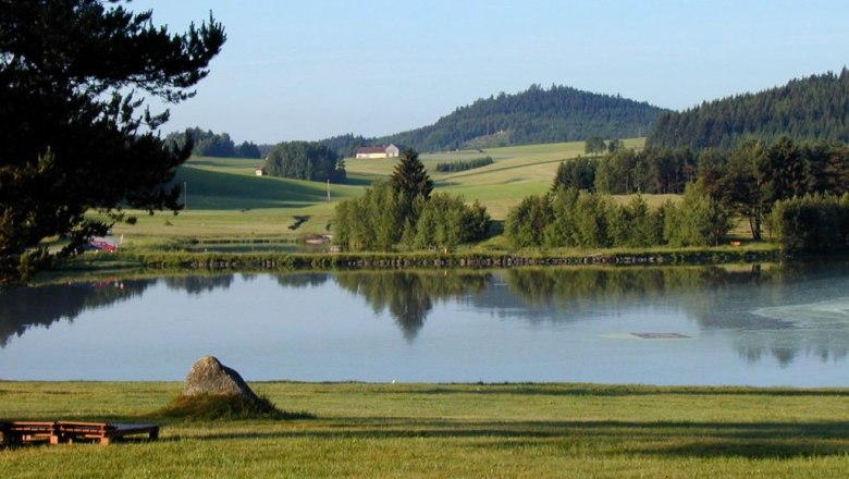 Ein ruhiger See mit umliegenden Wiesen und Wäldern in einer ländlichen Landschaft.