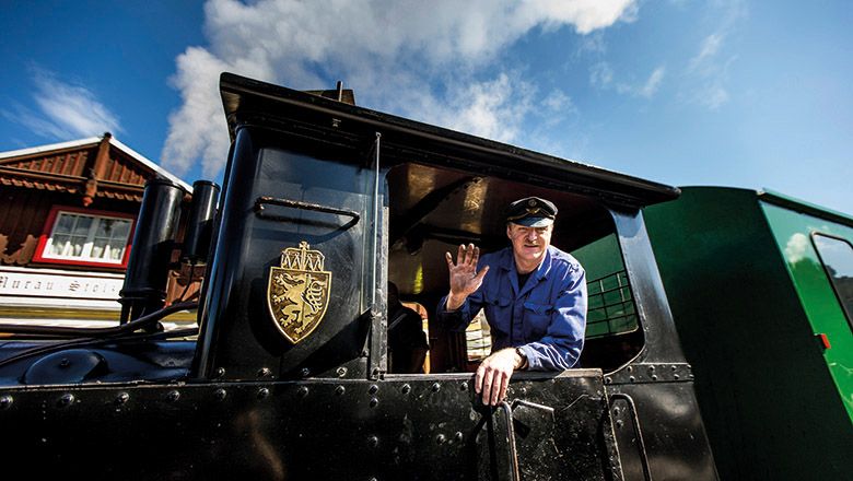 Lokführer in blauer Uniform lehnt aus dem Führerhaus einer schwarzen Dampflok der Murtalbahn und winkt. Im Hintergrund sind Bahnhof und blauer Himmel zu sehen.