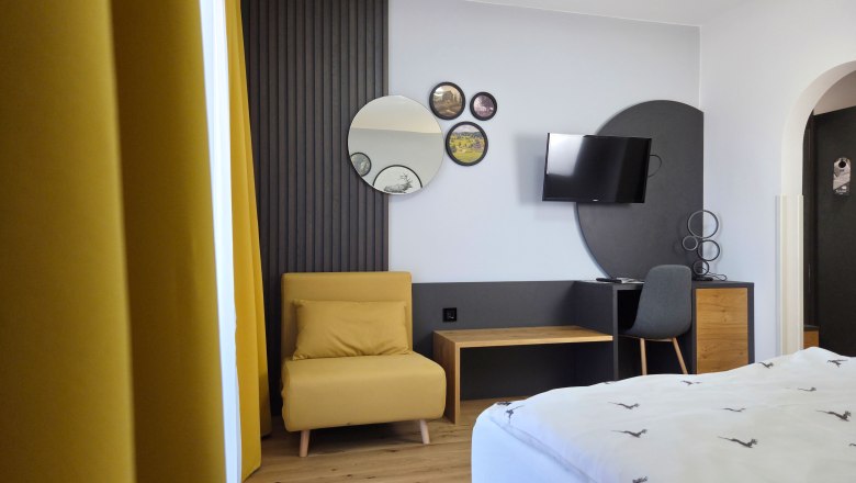 Modern eingerichtetes Zimmer mit gelbem Sessel, Spiegeln an der Wand, einem Fernseher und einem Schreibtisch mit Stuhl.