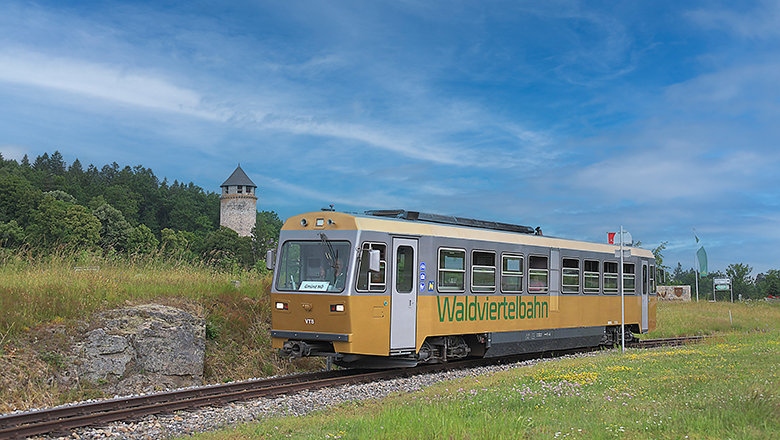 Ein goldener Zug der Waldviertelbahn f&auml;hrt durch eine gr&uuml;ne Landschaft mit einem Turm im Hintergrund.