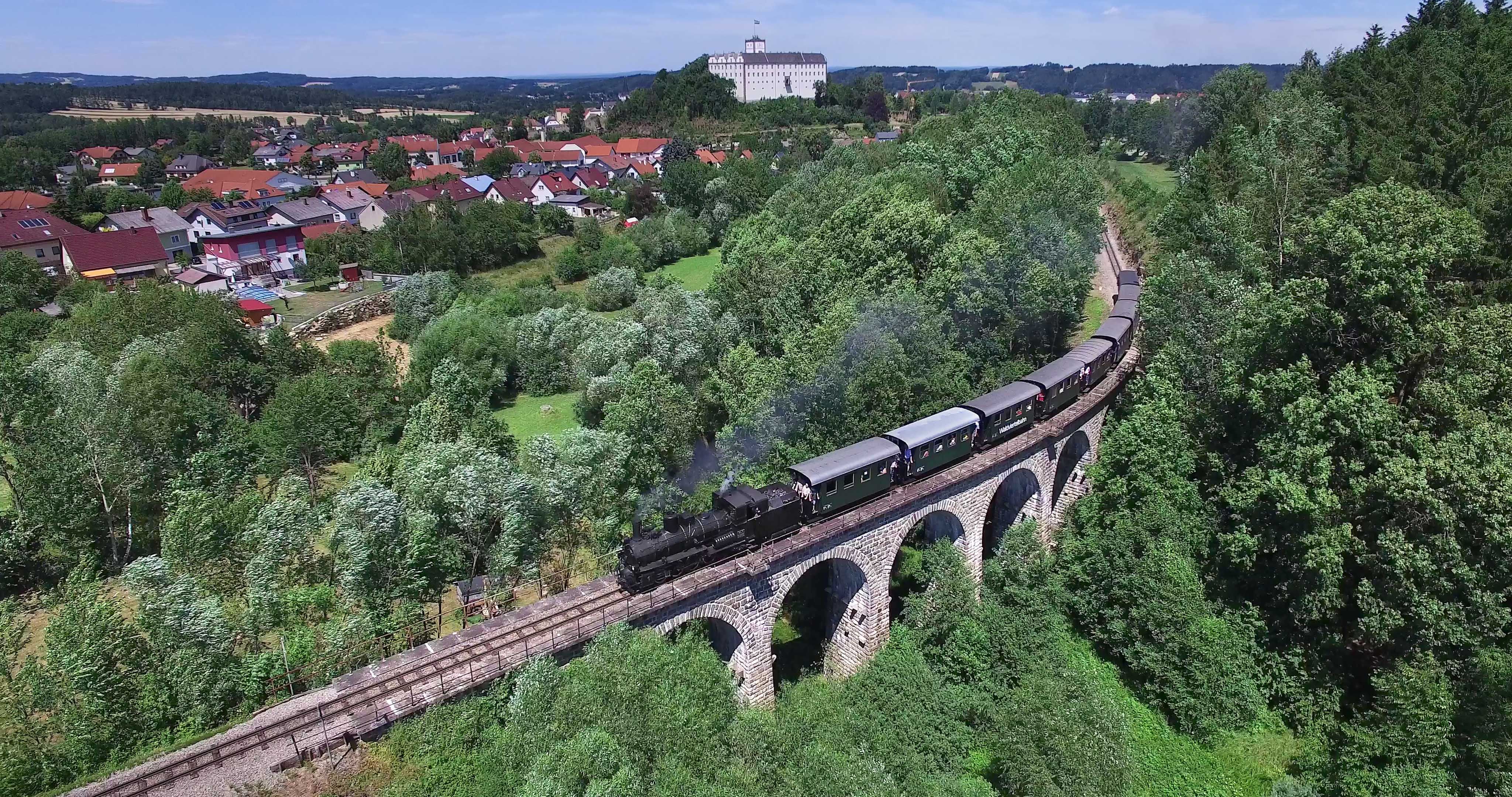Dampfzug mit grünen Waggons überquert einen steinernen Viadukt inmitten dichter Bäume. Im Hintergrund liegen Weitra und das Schloss.