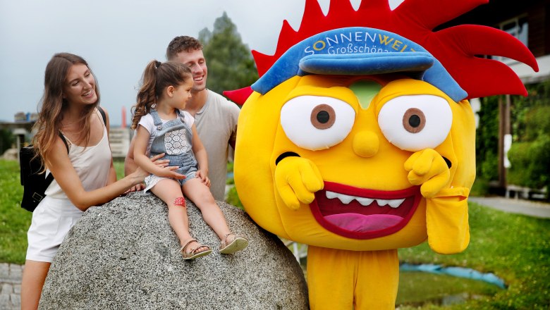 Familie mit Kind und Maskottchen auf einem Spielplatz.