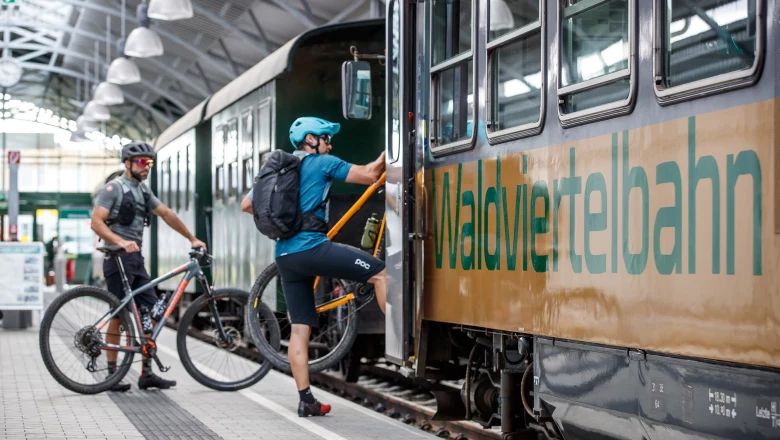 Eine Person lädt ein Fahrrad in einen Waggon der Waldviertelbahn, während eine zweite Person mit Fahrrad am Bahnsteig wartet.