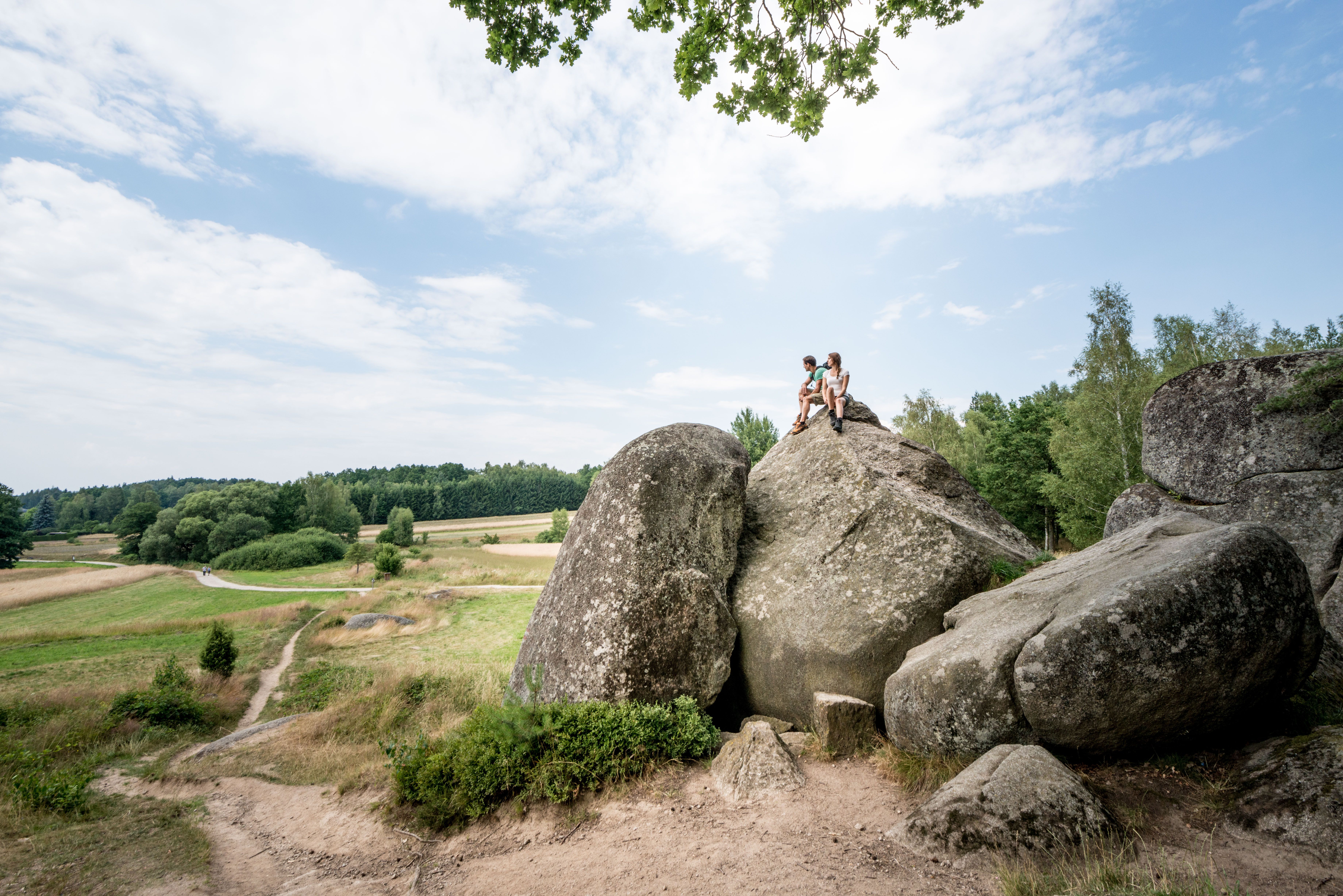 Zwei Personen sitzen auf einem großen Felsen in einer ländlichen Landschaft mit Wiesen und Bäumen.