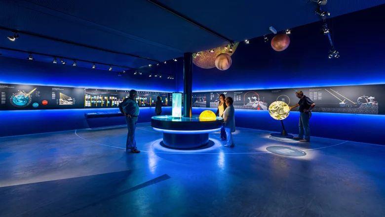 Blau beleuchteter Ausstellungsraum mit Personen an interaktiven Exponaten, Infotafeln an der Wand und Planetenmodellen an der Decke.