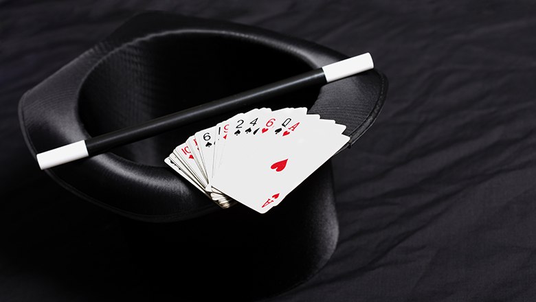 Ein schwarzer Zylinderhut mit einem Zauberstab und einem F&auml;cher aus Spielkarten darauf.