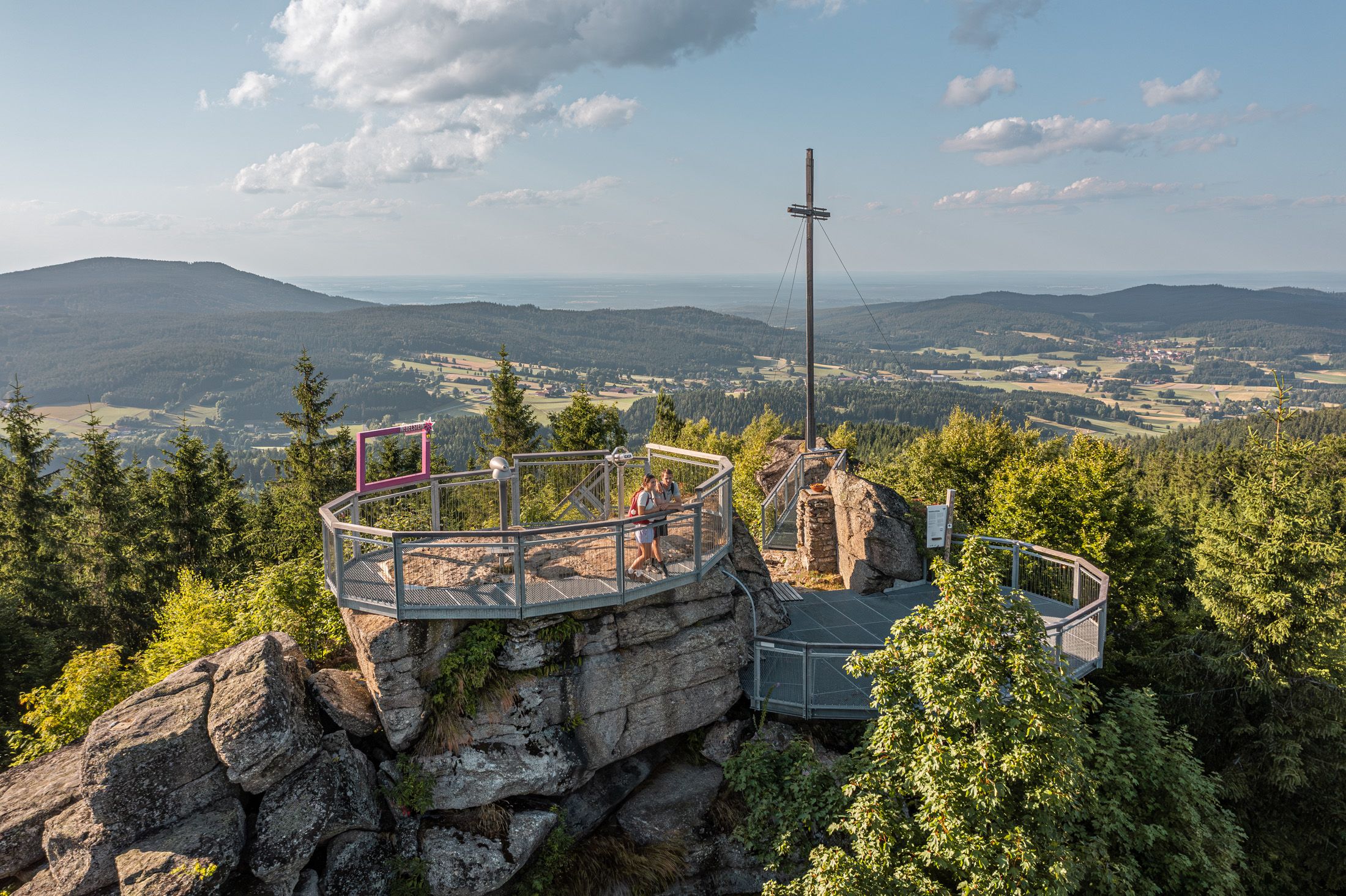 Aussichtsplattform auf einem Berg mit Kreuz und Blick auf bewaldete Landschaft.