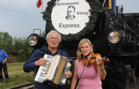 Zwei Musiker mit Akkordeon und Geige vor einer Dampflokomotive mit dem Schild 'Schrammel Klang Express'.