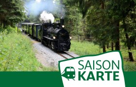 Dampflok f&auml;hrt durch einen Wald, begleitet von einem Schild mit der Aufschrift 'Saisonkarte'.