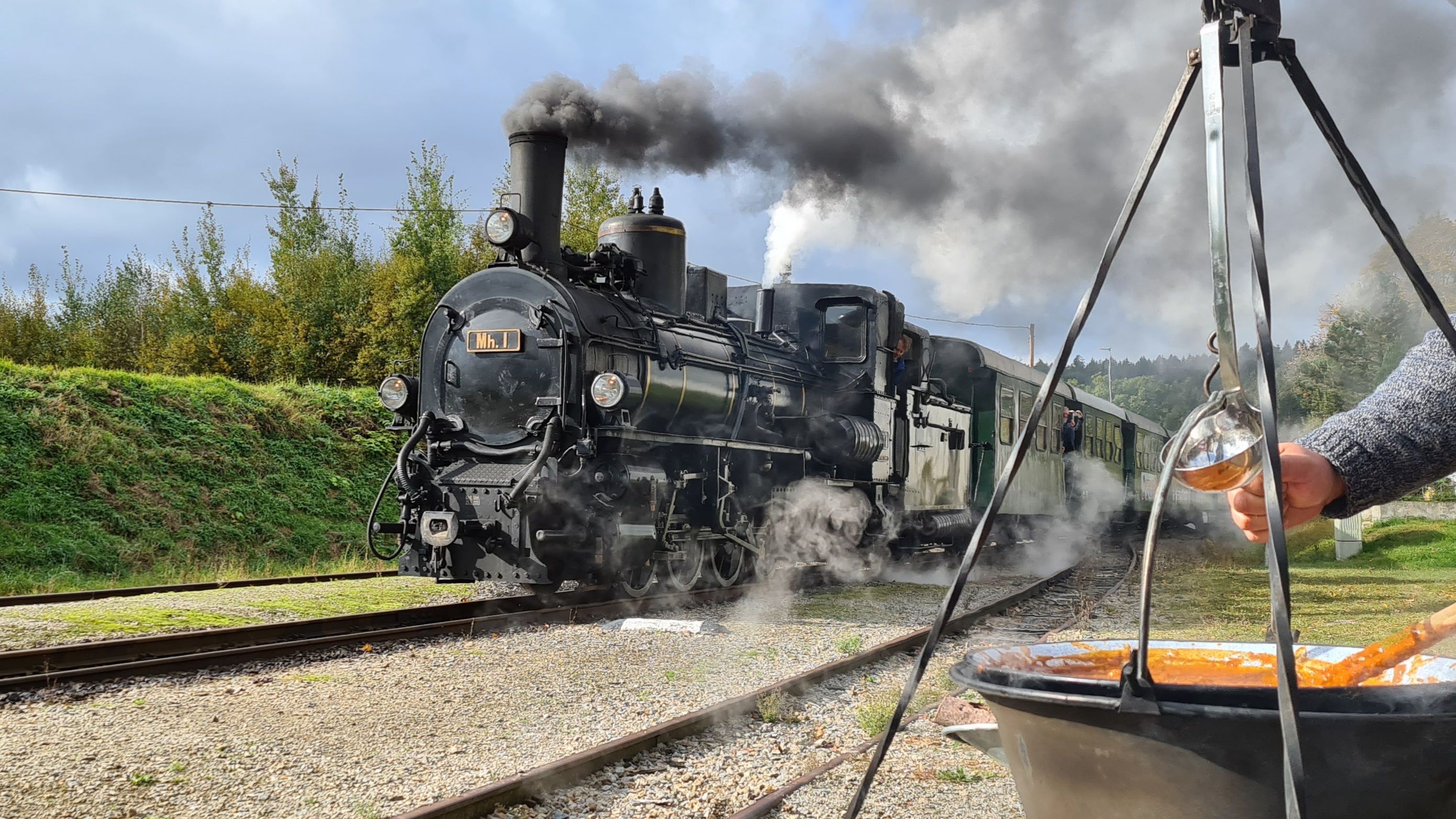 Eine Dampflokomotive fährt auf Schienen, während im Vordergrund ein Kessel mit Gulasch über einem Feuer hängt.
