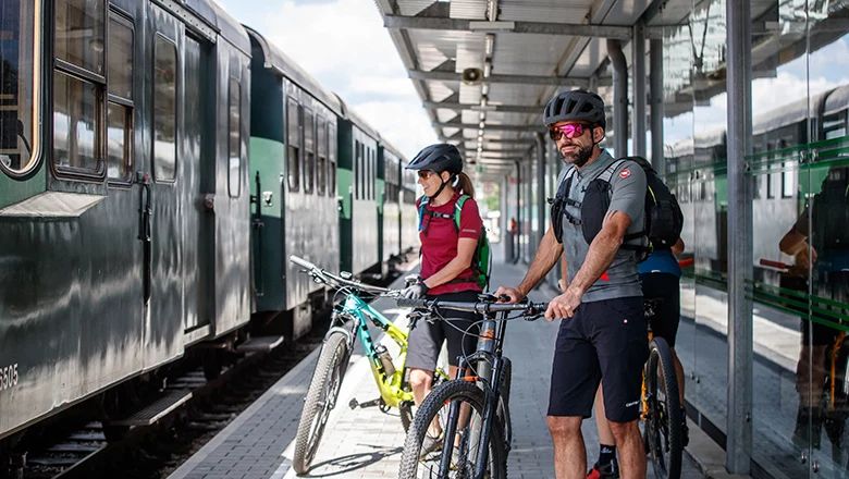 Zwei Personen stehen mit Mountainbikes auf einem Bahnsteig neben einem grünen Zug der Waldviertelbahn.