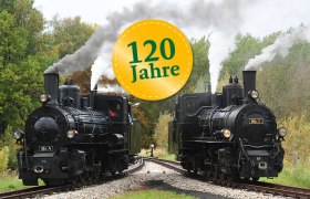 Zwei historische Dampflokomotiven auf einem Gleisdreieck in einer l&auml;ndlichen Umgebung mit einem Schild '120 Jahre'.