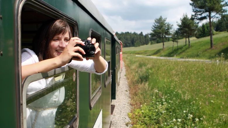Eine Frau lehnt sich aus dem Fenster eines grünen Zugwaggons und fotografiert die Wiesenlandschaft neben der Bahnstrecke.