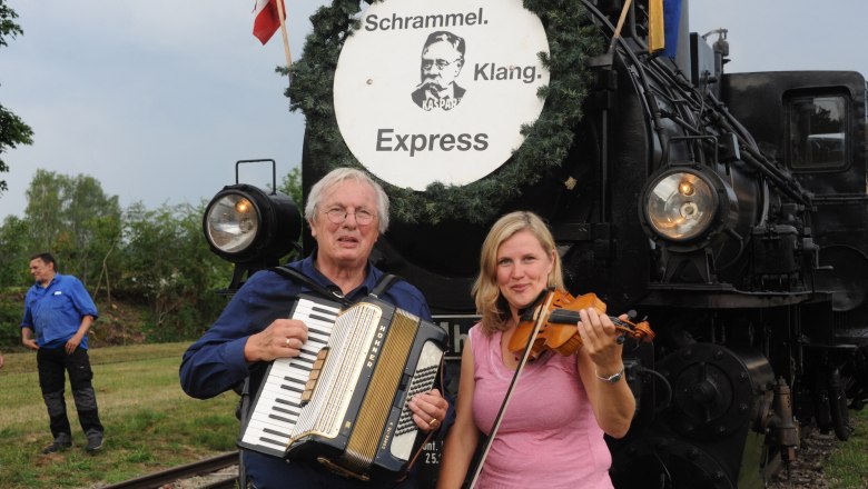 Zwei Musiker mit Akkordeon und Geige vor einer Dampflokomotive mit dem Schild 'Schrammel Klang Express'.
