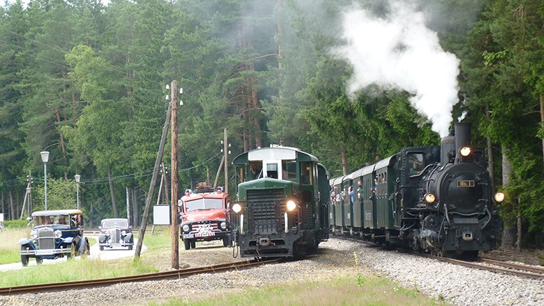 Ein Oldtimer-Auto, ein historischer Bus und eine Dampflokomotive auf einer l&auml;ndlichen Stra&szlig;e und Schiene.