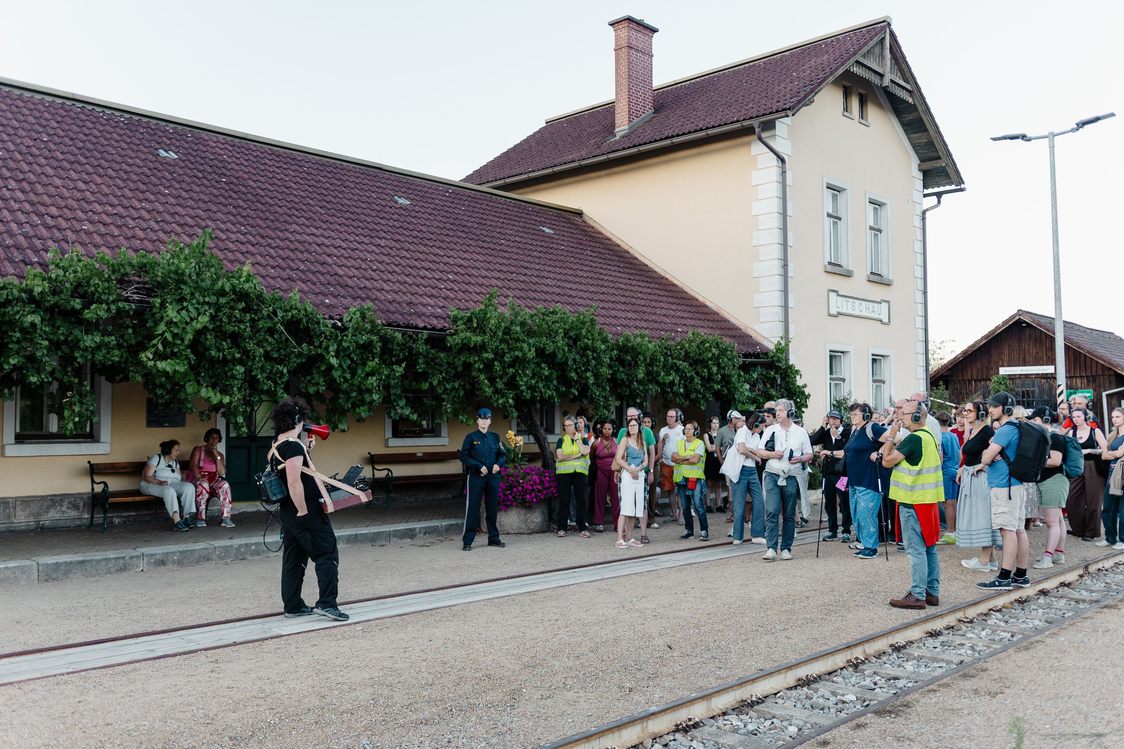 Menschen versammeln sich vor dem Gebäude Litschau an einem Bahnsteig; eine Person spricht mit Megafon, andere tragen Kopfhörer und Warnwesten.