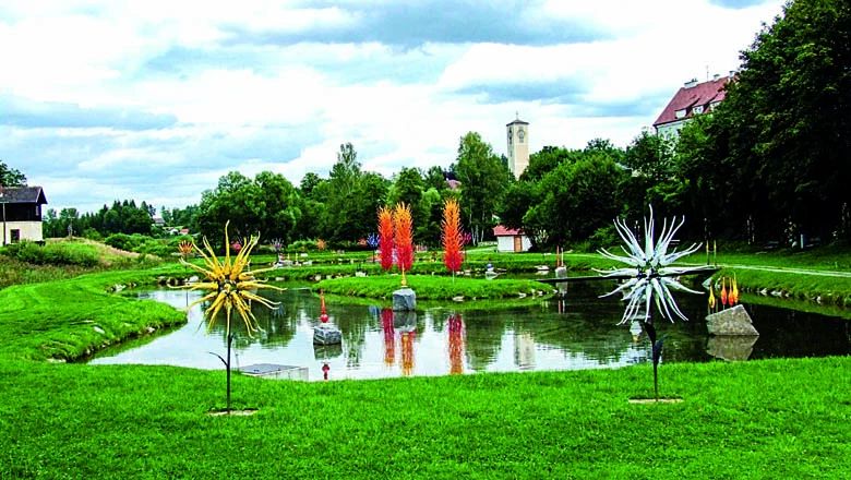 Bunte Glasskulpturen in Gelb, Orange und Weiß auf einer grünen Wiese und in einem Teich vor Bäumen und Gebäuden.