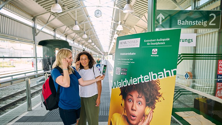 zwei Frauen stehen vor Werbeplakat für kostenlosen Audioguide